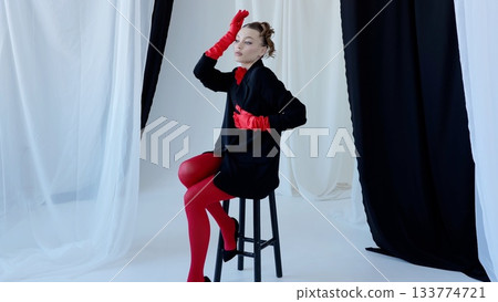 Fashion model posing sitting on a stool wearing red tights, red gloves and black oversized blazer in a studio with black and white curtains 133774721