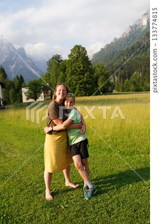 Mother and son share a joyful hug in a lush green field with mountain views. Their expressions are relaxed and happy, embodying a moment of connection and natural beauty. 133774815
