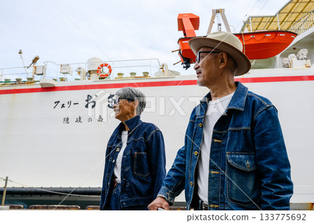 An elderly Japanese couple in denim jackets looks forward to the start of their trip at the port, with a ferry in the background 133775692