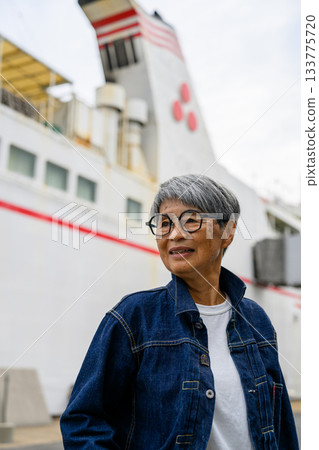 一位微笑的老婦人身穿牛仔外套，背景則是一艘渡輪。 133775720