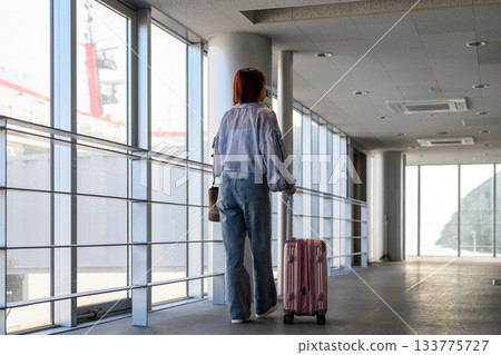 A young woman walking with a suitcase through a corridor with large windows 133775727