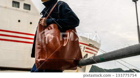 Man carrying a large brown leather tote bag with a ferry in the background at the port Man carrying a large brown leather tote bag with a ferry in the background at the port 133775754