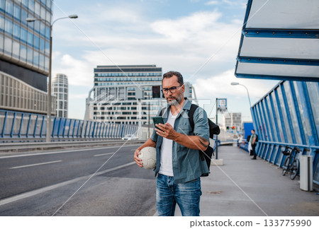 Bearded man with ball under arm using phone while waiting for bus. 133775990