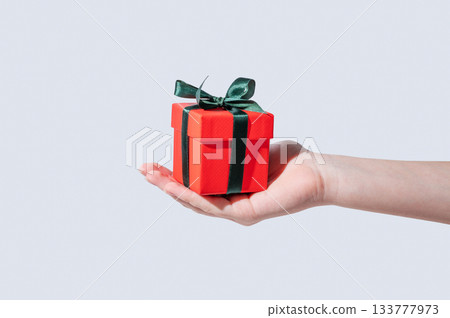 A hand holding a small red gift box with an emerald ribbon on an isolated light background A hand holding a small red gift box with an emerald ribbon on an isolated light background 133777973