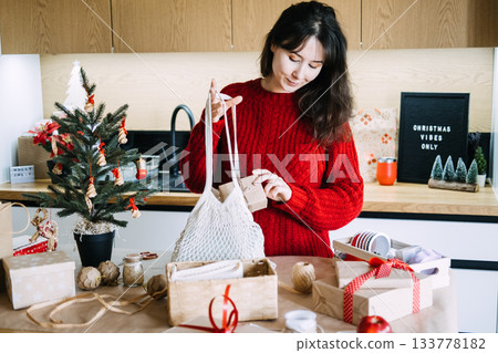 Young adult woman in red sweater places small gift box into reusable mesh bag on table with holiday supplies. Sustainable Self-Care Gift Ideas for Christmas, mindful gifting, eco wrapping 133778182