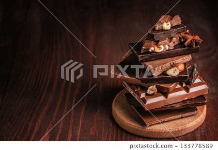 Assortment of stacked chocolate bars with nuts and spices on a wooden board placed on a dark rustic wooden background copy space, creating a warm and rich confectionery atmosphere for food Assortment of stacked chocolate bars with nuts and spices on a wooden board placed on a dark rustic wooden background copy space, creating a warm and rich confectionery atmosphere for food 133778589