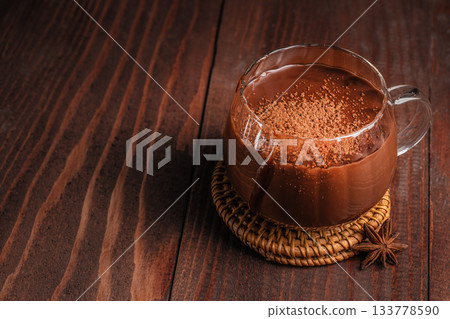 Rich hot chocolate in a clear glass mug topped with cocoa powder on a rustic wooden background copy space, creating a warm cozy dessert atmosphere perfect for winter drinks and sweet menu concepts Rich hot chocolate in a clear glass mug topped with cocoa powder on a rustic wooden background copy space, creating a warm cozy dessert atmosphere perfect for winter drinks and sweet menu concepts 133778590
