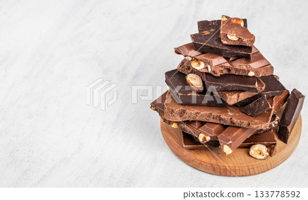 Assorted pieces of dark and milk chocolate with nuts stacked on light background copy space, perfect for dessert concepts, confectionery advertising and sweet food layouts 133778592