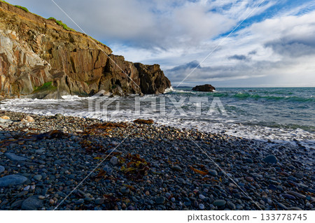 Pebbles and cliffs under a dynamic sky. Waves surge towards the shore. Pebbles and cliffs under a dynamic sky. Waves surge towards the shore. 133778745