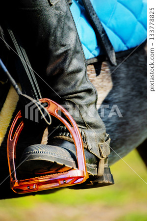 close-up of a brightly colored stirrup, with a rider's booted and cuisse-clad foot and a dressage whip in it. Photographed on a sunny day during equestrian training 133778825
