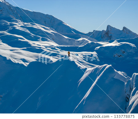 Aerial photo of a ski mountaineer standing on a snowy alpine ridge in Austria. Winter landscape, backcountry adventure, clear blue sky with sunshine and solitude in breathtaking mountain scenery. 133778875