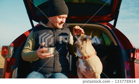 A Man Enjoying a Beautiful Day Outdoors with His Loyal Dog, Sharing Snacks and Moments of Joy from the Comfort of Their Vehicle's Open Trunk, Surrounded by Nature's Beauty 133778914