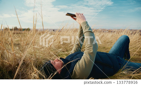 A Person Relaxing in a Natural Landscape, Lying on a Meadow, Capturing a Photo with a Camera Phone While Surrounded by Tall Grass Under a Clear Sky and Bright Afternoon Sunlight 133778916