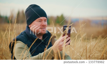 A man dressed in warm clothing sits in a field of tall grass while using a smartphone, enjoying a moment of connection with nature and technology in a tranquil outdoor setting. A man dressed in warm clothing sits in a field of tall grass while using a smartphone, enjoying a moment of connection with nature and technology in a tranquil outdoor setting. 133778982