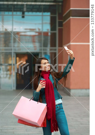 Beautiful Happy Woman Holding Shopping Bags And Smiling Near The Mall. Female taking selfie photos at smartphone. 133779148