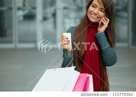 Young woman with shopping bags walking out from shop and talking on the phone. Woman with coffee. Consumerism, shopping Young woman with shopping bags walking out from shop and talking on the phone. Woman with coffee. Consumerism, shopping 133779362