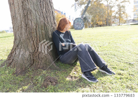 Young woman enjoying book outdoors leaning against tree Young woman enjoying book outdoors leaning against tree 133779683