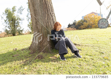 Woman reading book leaning against large tree Woman reading book leaning against large tree 133779685