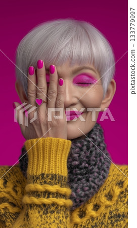 Vibrant, close-up portrait of an elderly Asian woman with stylish silver hair, hiding one eye with a hand featuring a pink heart ring, bold pink makeup, and a yellow sweater. 133779997