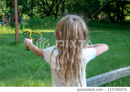 Child practices aiming with a slingshot in a green backyard during a sunny afternoon 133780078