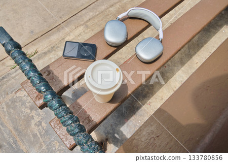 Headphones, music player, coffee cup on park bench Headphones, music player, coffee cup on park bench 133780856