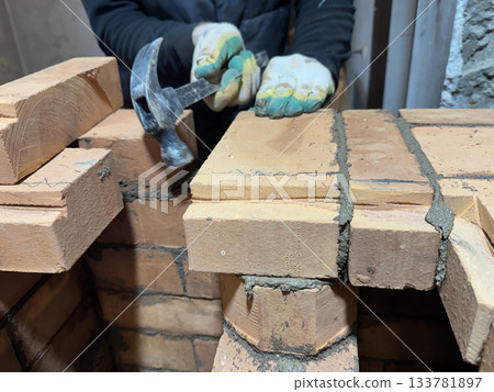 A stove fitter adjusts a laid brick using a hammer. Brickwork of a heating stove. Channels for dividing and passing smoke. Close-up. A stove fitter adjusts a laid brick using a hammer. Brickwork of a heating stove. Channels for dividing and passing smoke. Close-up. 133781897