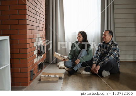 Adding firewood to heat stove by caucasian man and woman sitting together on floor in warm indoor rustic room with natural light. Winter Firewood Routine Preparing and Lighting a Wood Stove 133782100