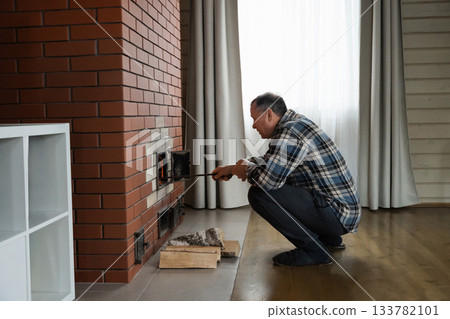 Caucasian adult man tending flames with poker inside burning brick stove while heating indoor room in rustic warm atmosphere. Winter Firewood Routine Preparing and Lighting a Wood Stove 133782101