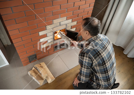 Caucasian adult man adding large log into burning brick stove while warming indoor room during cold season. Cold season. Winter Firewood Routine Preparing and Lighting Wood Stove Caucasian adult man adding large log into burning brick stove while warming indoor room during cold season. Cold season. Winter Firewood Routine Preparing and Lighting Wood Stove 133782102