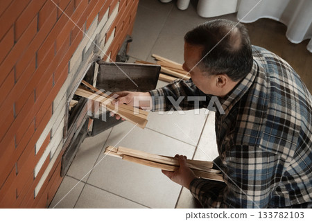 Placing thin kindling into brick stove by caucasian adult man in checkered shirt while preparing fire inside warm indoor room. Winter Firewood Routine Preparing and Lighting Wood Stove Placing thin kindling into brick stove by caucasian adult man in checkered shirt while preparing fire inside warm indoor room. Winter Firewood Routine Preparing and Lighting Wood Stove 133782103