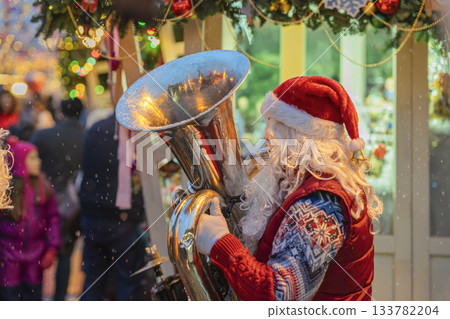 Santa Claus playing tuba at outdoor Christmas market with festive decorations and falling snow. Concept of holiday celebration, music and joyful spirit of Christmas tradition 133782204