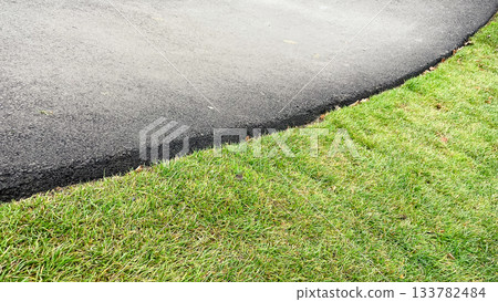 Sharp dividing line between lush green lawn and smooth grey asphalt road surface. Abstract texture contrast Sharp dividing line between lush green lawn and smooth grey asphalt road surface. Abstract texture contrast 133782484