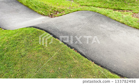 Curved asphalt pump track for cycling and wheeled sports, surrounded by vibrant green grass in a modern park Curved asphalt pump track for cycling and wheeled sports, surrounded by vibrant green grass in a modern park 133782498