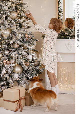 Caucasian blonde girl decorating snowy Christmas tree with corgi in cozy home interior Caucasian blonde girl decorating snowy Christmas tree with corgi in cozy home interior 133782984