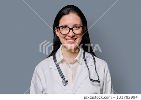 Portrait of smiling female doctor in white lab coat with stethoscope on gray background Portrait of smiling female doctor in white lab coat with stethoscope on gray background 133782994