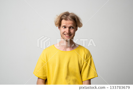 Smiling young man looking sideways with awkward amused expression while posing in neutral studio scene 133783679