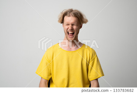 Young man in yellow shirt screaming loudly with eyes closed and emotional expression in studio portrait 133783682