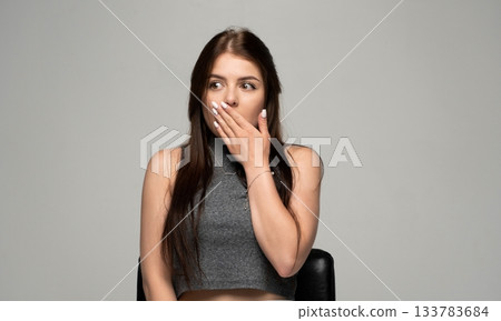 Surprised young woman covering her mouth with hand while looking sideways in studio on grey background Surprised young woman covering her mouth with hand while looking sideways in studio on grey background 133783684