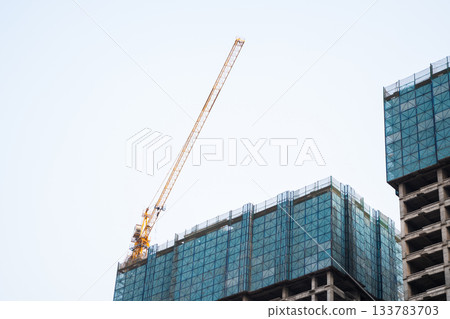 Modern building under construction with glass panels and high tower crane against blue sky Modern building under construction with glass panels and high tower crane against blue sky 133783703