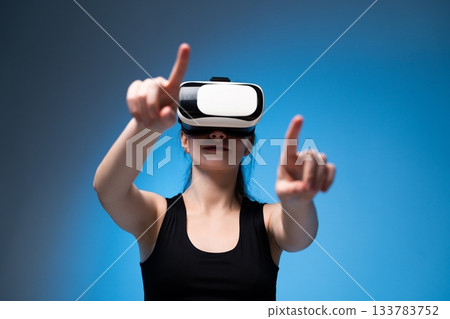 Woman in VR headset pointing at virtual elements while interacting with digital environment on blue background 133783752