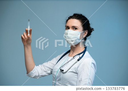 Nurse holding syringe upright preparing medical injection while wearing protective mask in clinical setting Nurse holding syringe upright preparing medical injection while wearing protective mask in clinical setting 133783776