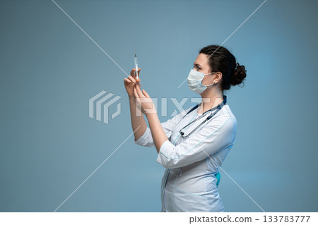 Nurse examining syringe before injection while wearing mask and uniform in blue clinical background Nurse examining syringe before injection while wearing mask and uniform in blue clinical background 133783777
