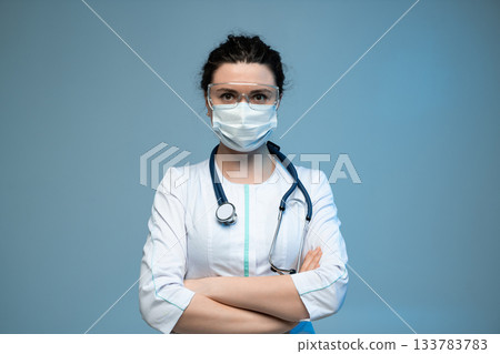 Female doctor with crossed arms wearing mask and protective glasses standing on blue medical background 133783783