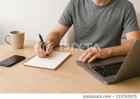 The man is working on a laptop and writing notes. The man is working on a laptop and writing notes. 133783970