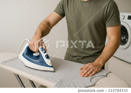 A man irons a gray T-shirt with an iron. 133784335