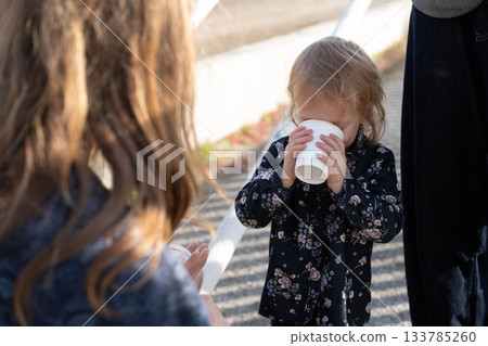 A young child savors a warm beverage while surrounded by family members during a lovely outdoor gathering. The setting is peaceful, showcasing the beauty of nature. 133785260