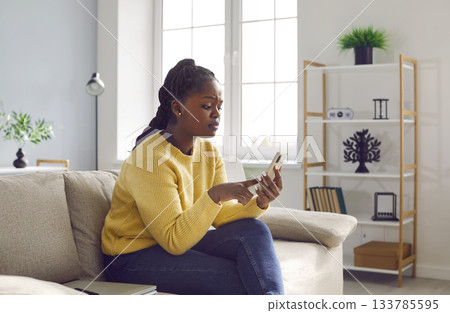 Young african girl with worried face expression using mobile phone while sitting on sofa at home. 133785595