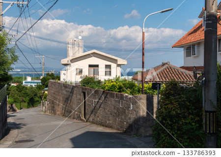 A hilly backstreet in the countryside on a sunny day with a view of Okinawa's red tiled roofs and the emerald green ocean in the distance A hilly backstreet in the countryside on a sunny day with a view of Okinawa's red tiled roofs and the emerald green ocean in the distance 133786393