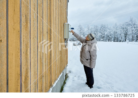 Mature woman checks electricity meters mounted on yellow wooden siding house wall. 133786725