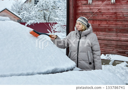 Brushing removes deep snow from car roof as woman in warm winter jacket and knit hat clears vehicle near wooden wall in snowy yard. 133786741